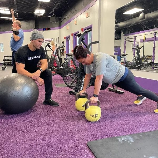 Woman performing a kettlebell plank exercise while a personal trainer supervises in a gym