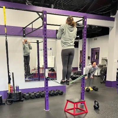 Woman performing an assisted pull-up using a resistance band in a gym while another person encourages her